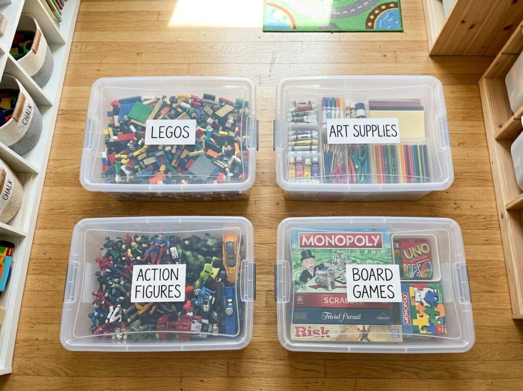 Top-down view of clear plastic storage bins neatly organized on a playroom floor with printed labels for legos, art supplies, and board games.