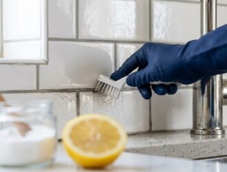 A close-up photograph of a gloved hand using a white brush to scrub the gray grout lines between white subway tiles behind a kitchen faucet, next to a blurred lemon and baking soda.
