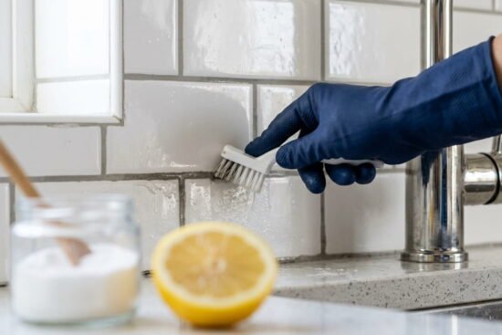 A close-up photograph of a gloved hand using a white brush to scrub the gray grout lines between white subway tiles behind a kitchen faucet, next to a blurred lemon and baking soda.