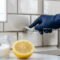 A close-up photograph of a gloved hand using a white brush to scrub the gray grout lines between white subway tiles behind a kitchen faucet, next to a blurred lemon and baking soda.