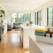 A wide-angle photograph of a pristine, sunlit kitchen and living area with white cabinets and hardwood floors, gleaming after a complete deep clean. A cleaning caddy stocked with microfiber cloths, spray bottles, and a grout brush sits on the kitchen island.