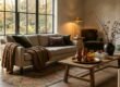 A sunlit, cozy living room styled for autumn with a cream sofa, camel bouclé throw, moss green velvet pillows, aged brass decor, and tall branches in a ceramic urn.